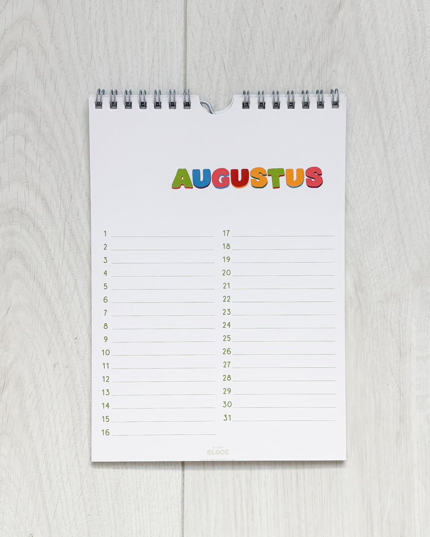 Verjaardagskalender op A5-formaat met ringband, crèmekleurige basis en kleurrijke handgetekende letters per maand; veel schrijfruimte en minimalistisch ontwerp van Studio Bloos – opgehangen aan de muur.