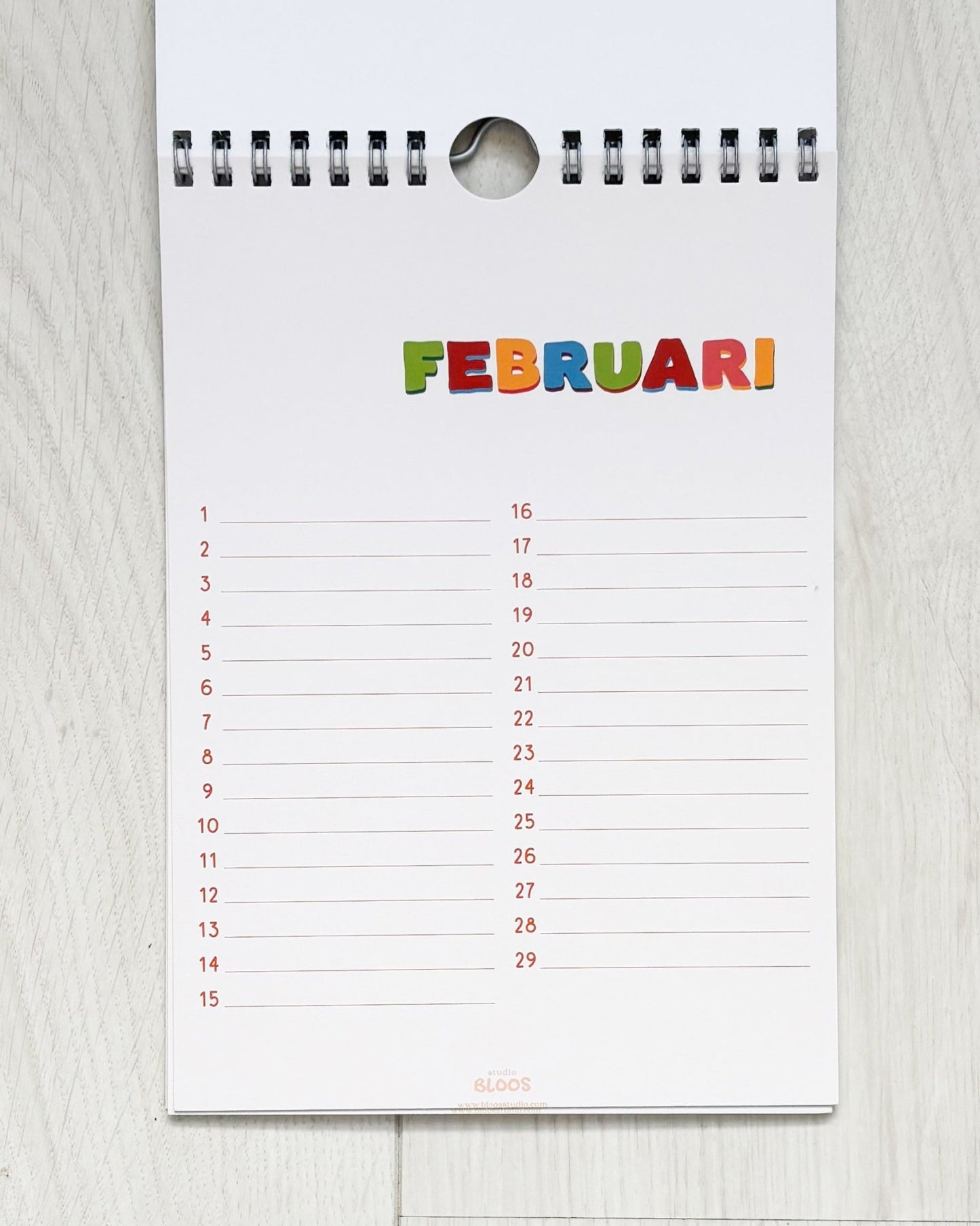 Verjaardagskalender op A5-formaat met ringband, crèmekleurige basis en kleurrijke handgetekende letters per maand; veel schrijfruimte en minimalistisch ontwerp van Studio Bloos – opgehangen aan de muur.
