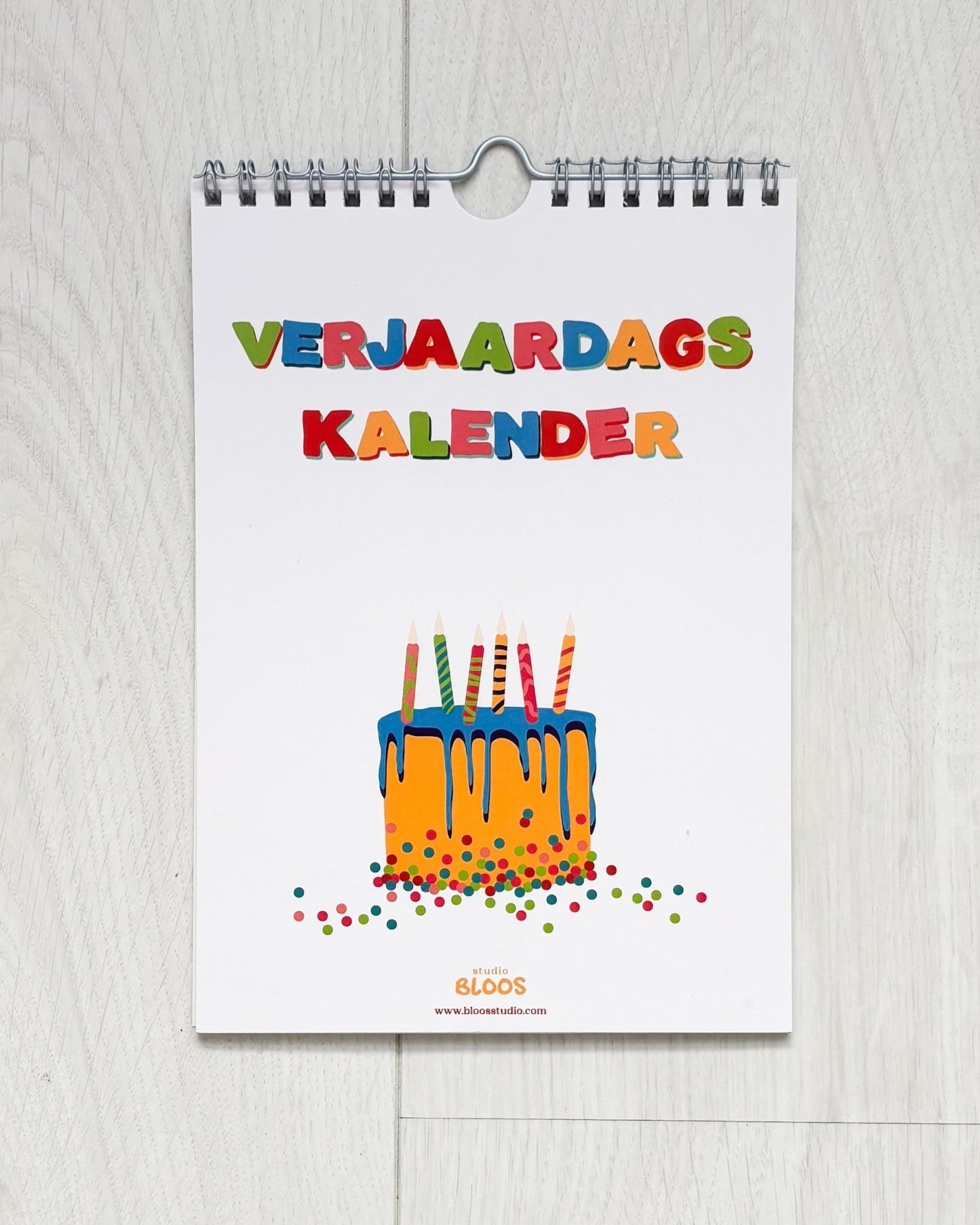 Verjaardagskalender op A5-formaat met ringband, crèmekleurige basis en kleurrijke handgetekende letters per maand; veel schrijfruimte en minimalistisch ontwerp van Studio Bloos – opgehangen aan de muur.