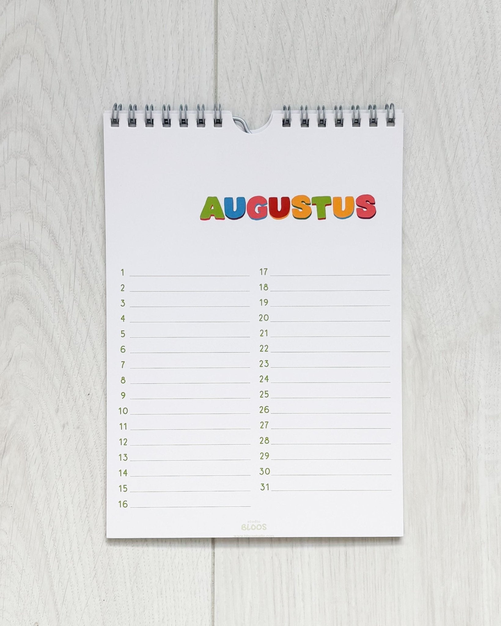 Verjaardagskalender op A5-formaat met ringband, crèmekleurige basis en kleurrijke handgetekende letters per maand; veel schrijfruimte en minimalistisch ontwerp van Studio Bloos – opgehangen aan de muur.