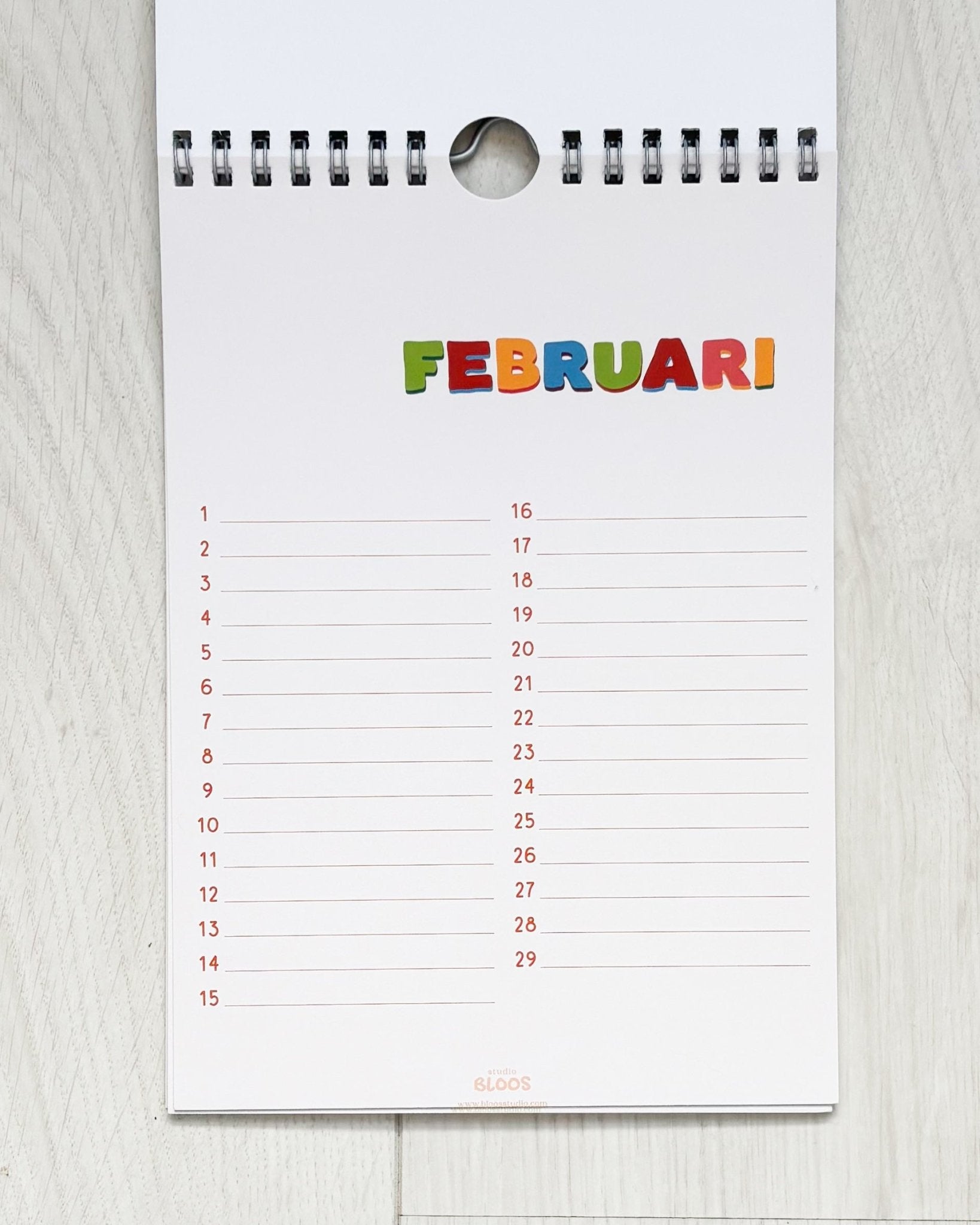 Verjaardagskalender op A5-formaat met ringband, crèmekleurige basis en kleurrijke handgetekende letters per maand; veel schrijfruimte en minimalistisch ontwerp van Studio Bloos – opgehangen aan de muur.