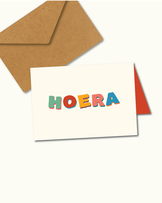 Wenskaart met kleurrijke handgetekende letters “Hoera” op een crèmekleurige achtergrond – feestelijk ontwerp van Studio Bloos.