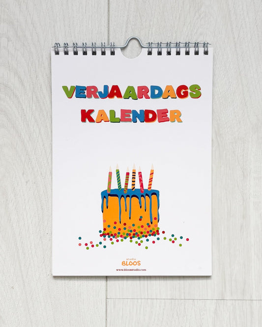 Verjaardagskalender op A5-formaat met ringband, crèmekleurige basis en kleurrijke handgetekende letters per maand; veel schrijfruimte en minimalistisch ontwerp van Studio Bloos – opgehangen aan de muur.